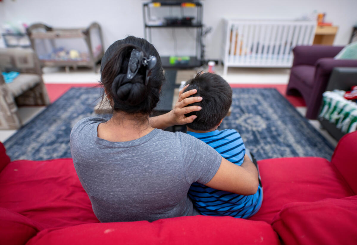 A mother sits on a red couch with her arm around her son sitting next to her. They are in a room with several chairs and a large rug. 