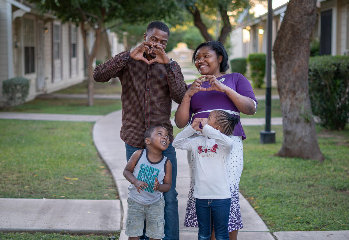 A couple and two children pose outside a row of houses and making a heart symbol with their hands.