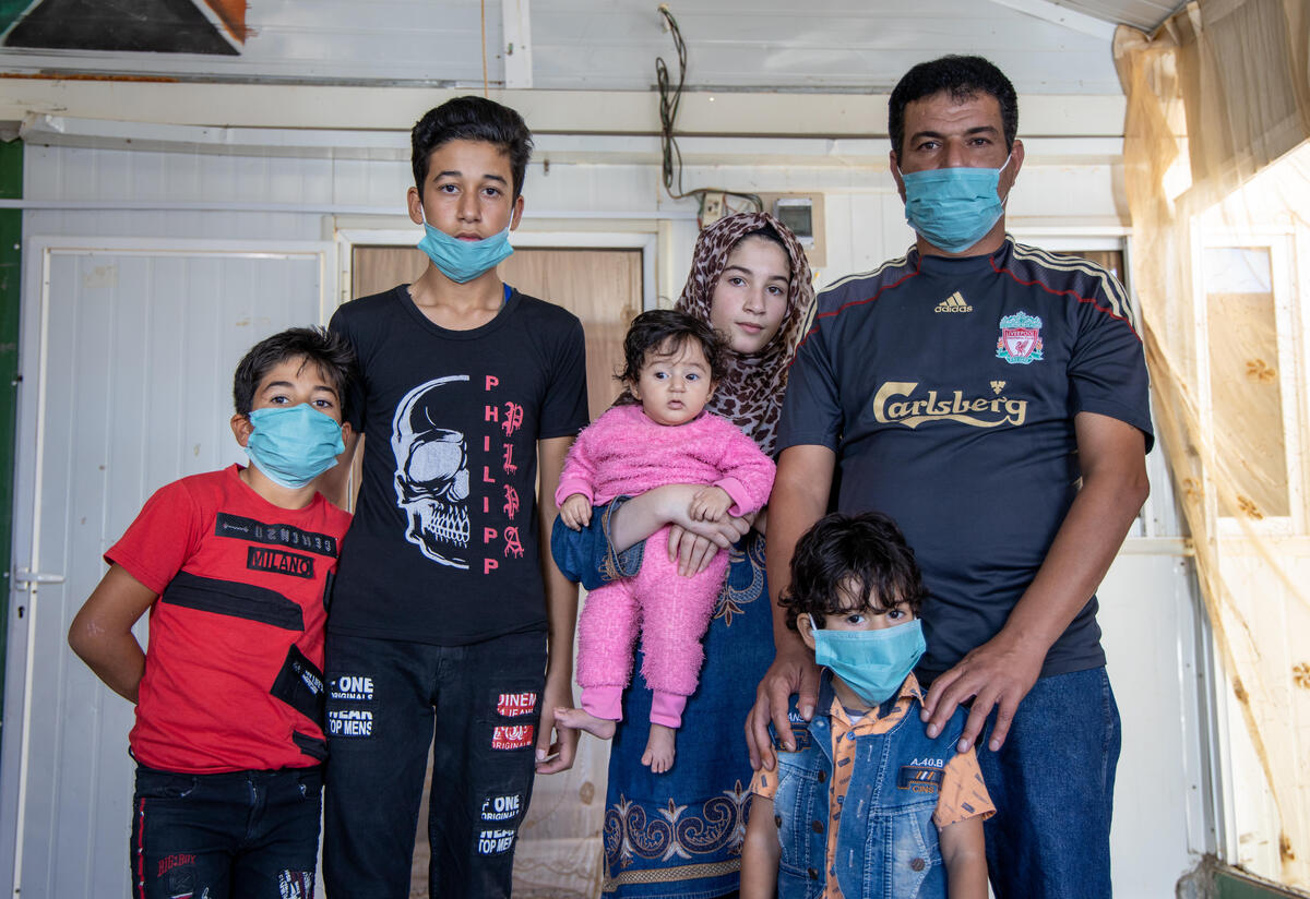 Manhal and his family In a small tin home in a refugee camp, Manhal poses with his family: his wife holds a baby in a pink onesie, his four-year-old stands in front of him and eight-year-old and fifteen-year-old sons stands next to them.