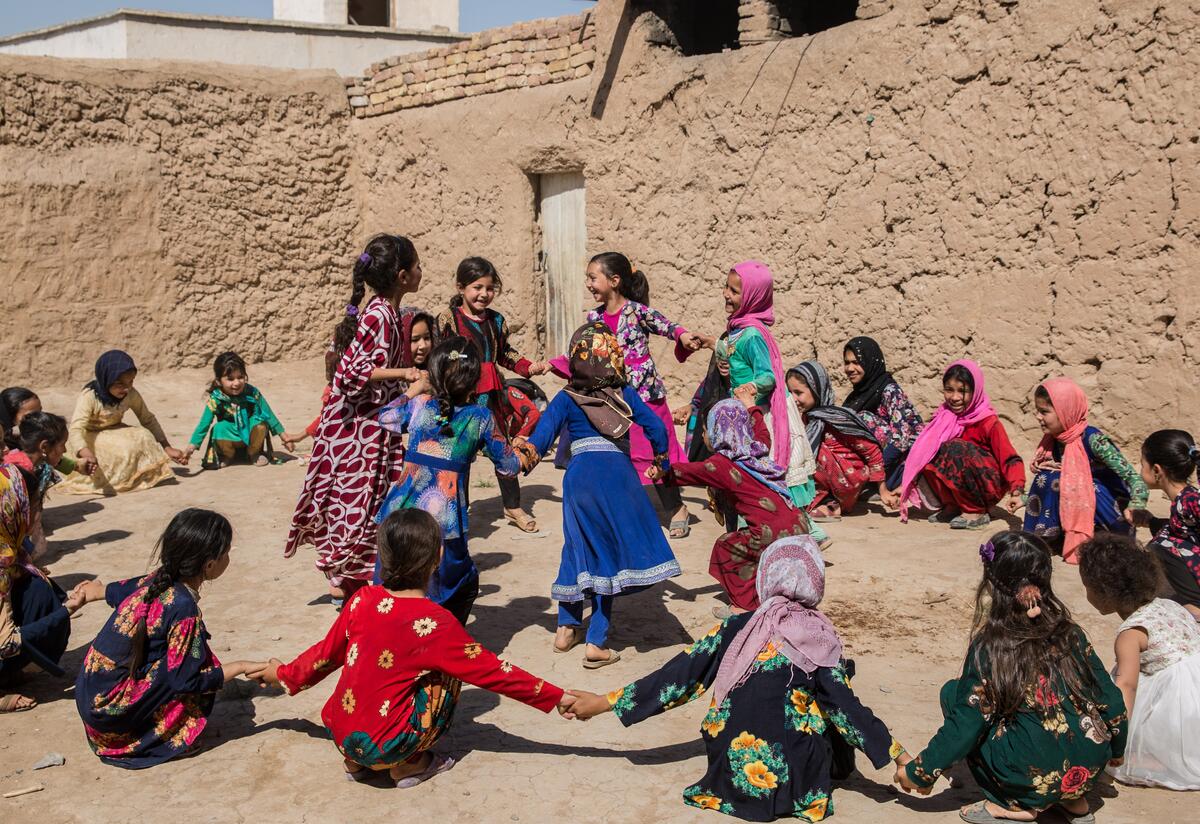 Young schoolgirls in Afghanistan hold hands sitting in a ring around a group of classmaters who are dancing.