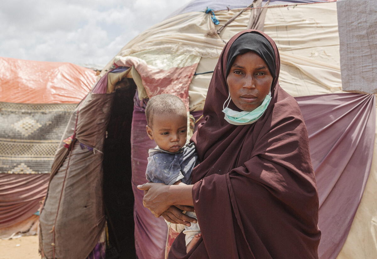 Amina with her son. Amina stands in front of tents outside while holding their son.