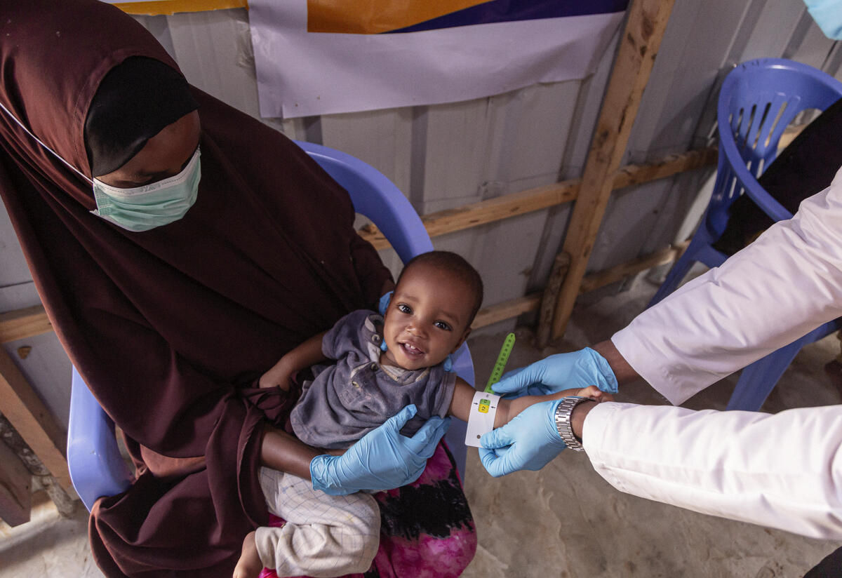 Amina holds her son while an IRC staff member wearing a white coat and blue gloves measures his arm with a color-coded armband. 