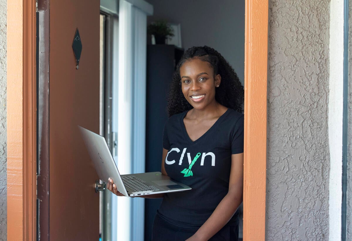 Diana Muturia stands in a doorway in her home holding her laptop and wearing a shirt with the name of her app, "Clyn" 