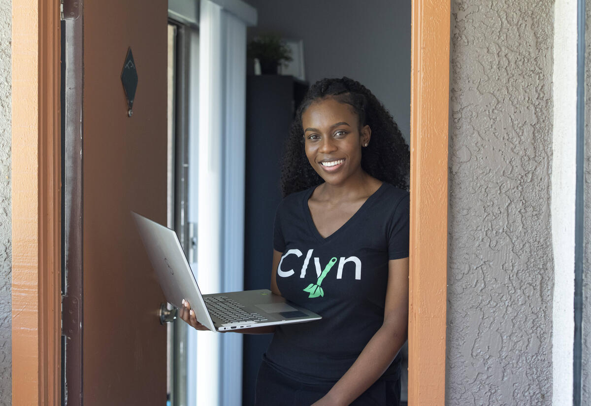 Diana Muturia Diana Muturia stands in a doorway in her home holding her laptop and wearing a shirt with the name of her app, "Clyn"