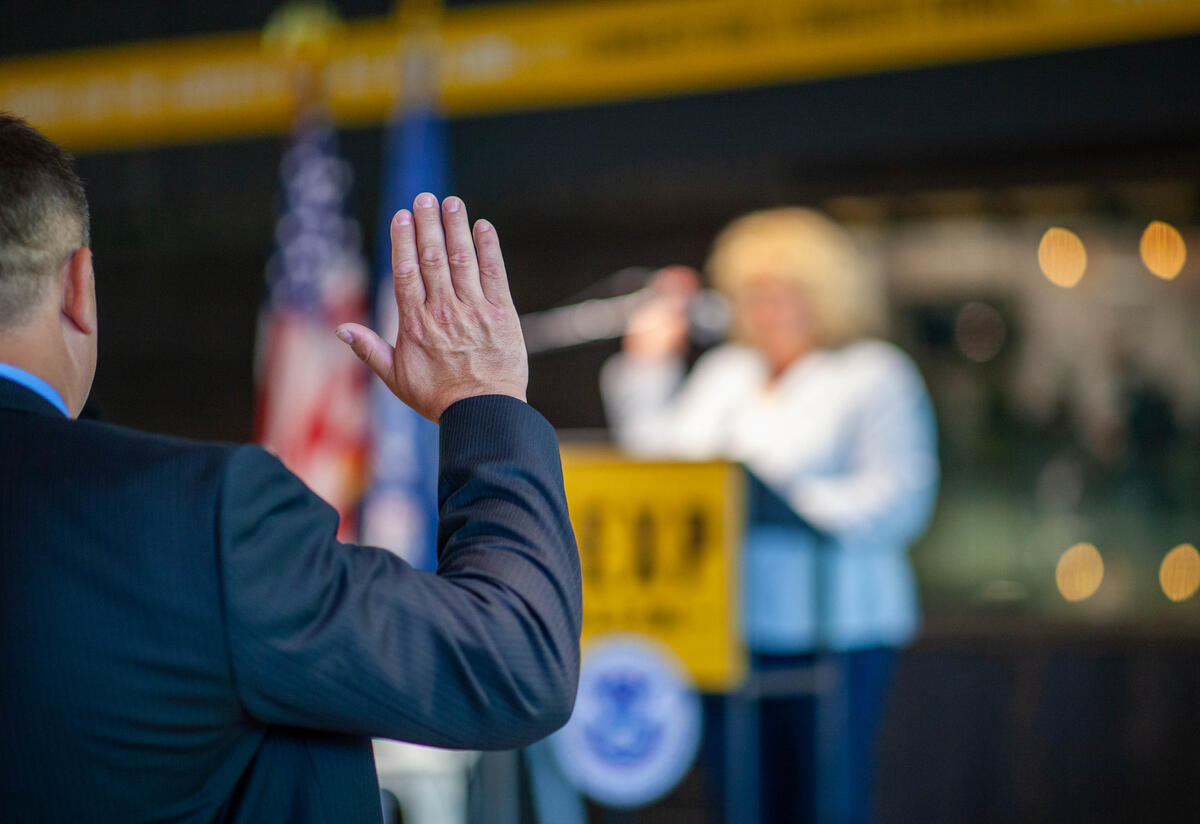 Man wearing a suit, raising hand and taking oath during naturalization ceremony. A view of man from the back raising his hand, taking an oath during a naturalization ceremony.