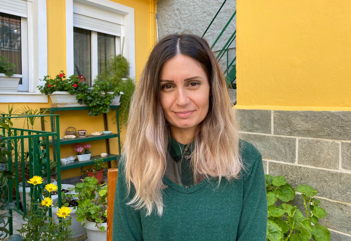 Kiki Michailidou, wearing a green sweater, sits outside a yellow house near a garden