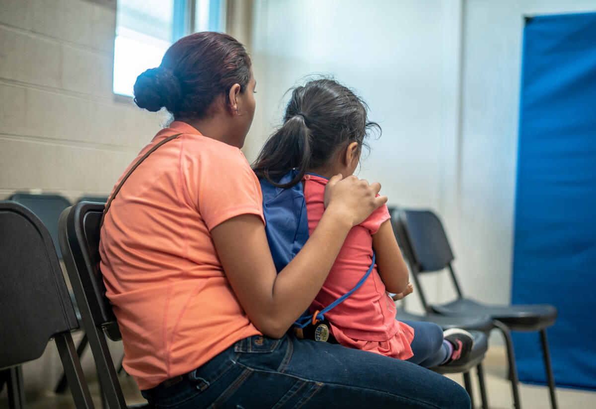 In an IRC shelter, a young mother sits with her back to the camera and her arm around her 4-year-old daughter. 