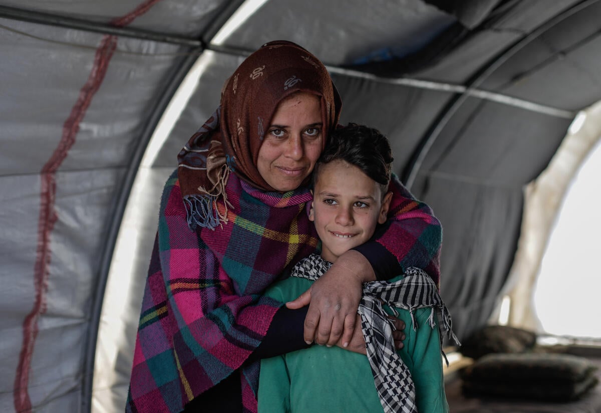 Sundus hugs her son Omar while standing behind him in a tent. Both are smiling and looking at the camera. 