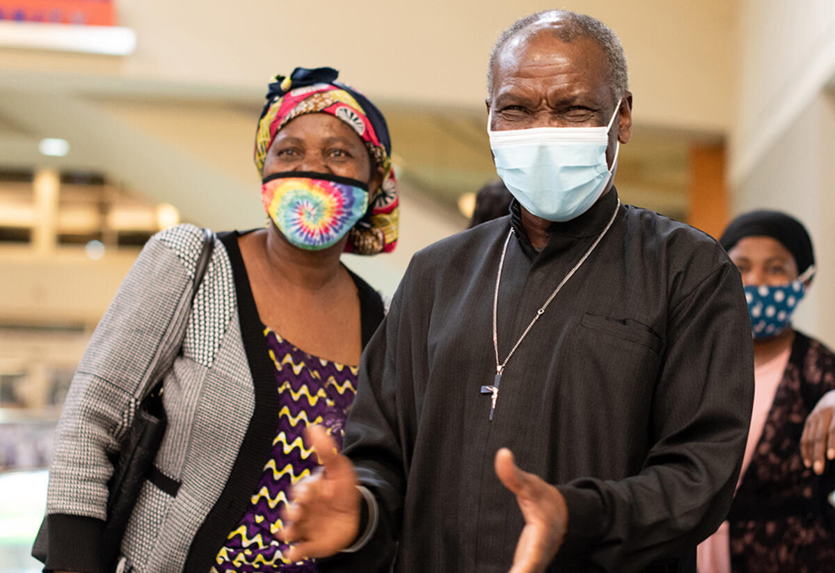 Patrice and Wanyema, both wearing masks, both watch their daughter arrive at the Boise airport. Patrice is about to clap his hands. 