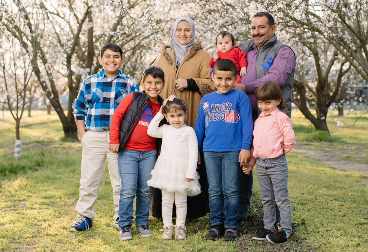 A husband and wife from Syria and their six children are standing and smiling in the middle of an almond orchard when the almond trees are blooming.