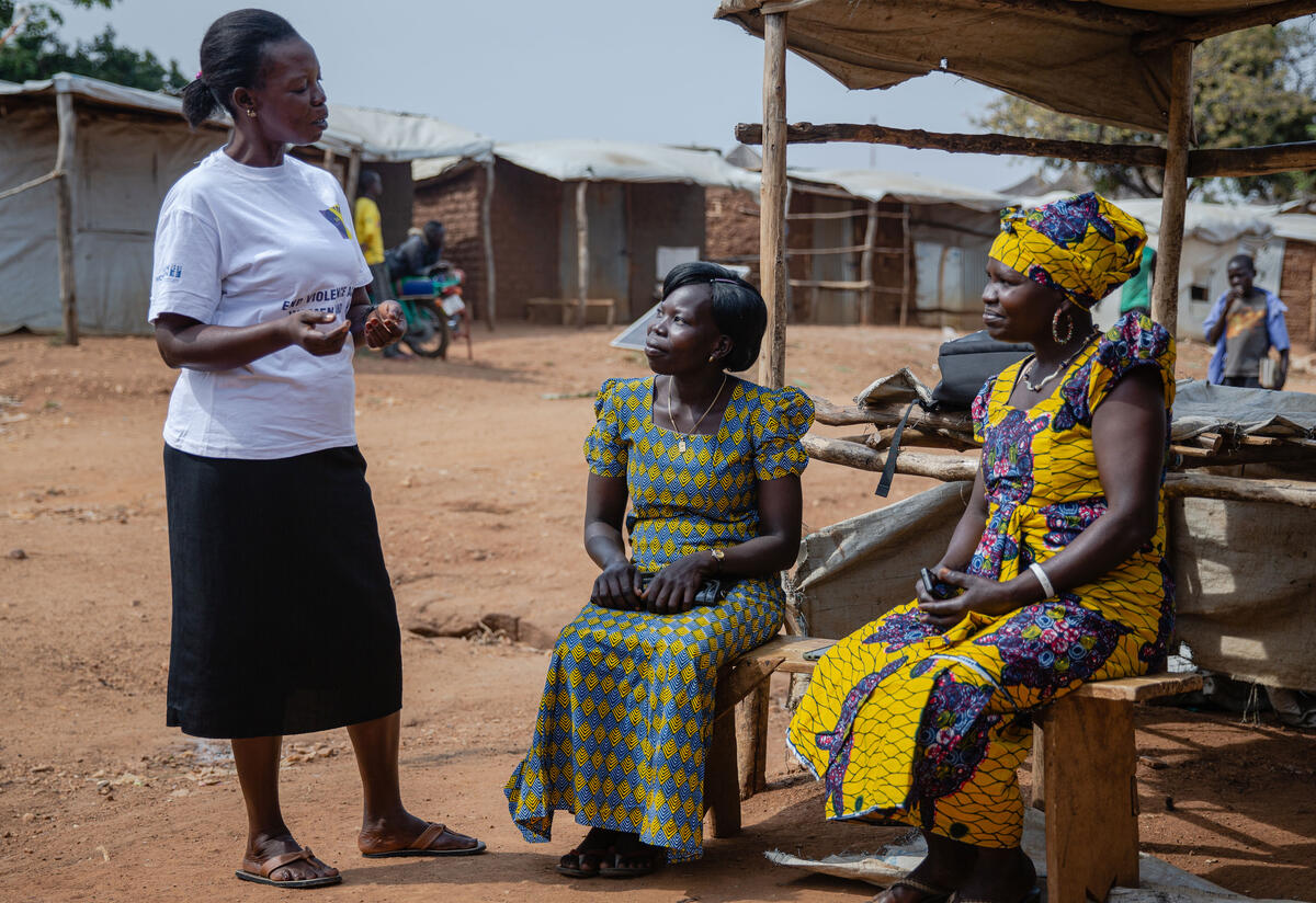 Three women talk to one another in a refugee camp, two sitting down and the other standing. 