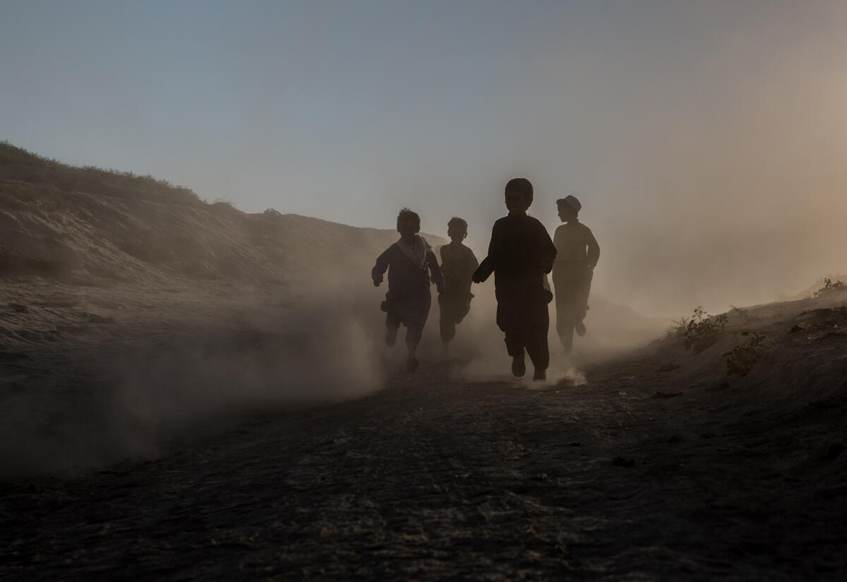 Children run through a camp in Afghanistan for people displaced by drought. Children run through a desert and mountain landscape. The light is such that their faces are obscured.