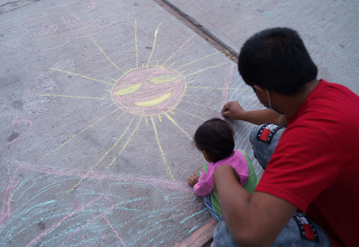 A parent and child draw with chalk at the triage hotel. A father and young child bend over the ground drawing with chalk. They have already drawn a large sun.