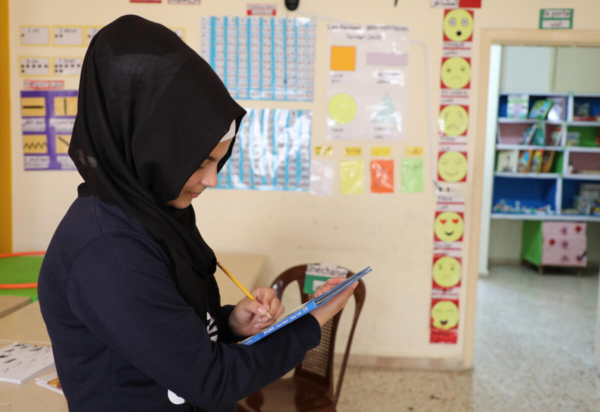 Widad, a young Syrian refugee, at an IRC-run safe space in northern Lebanon A young girl stands and writes in a book she is holding. She is in a brightly-colored room full of educational materials.