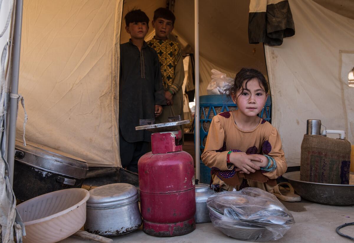 A displaced Afghan girl crouches outside her tent in Afghanistan with two of her brothers standing behind her in the doorway.