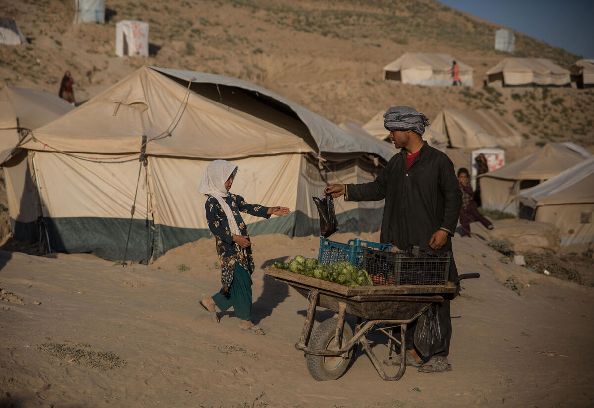 With tents in the background, a man with a fruit cart hands a bag to a young girl. 