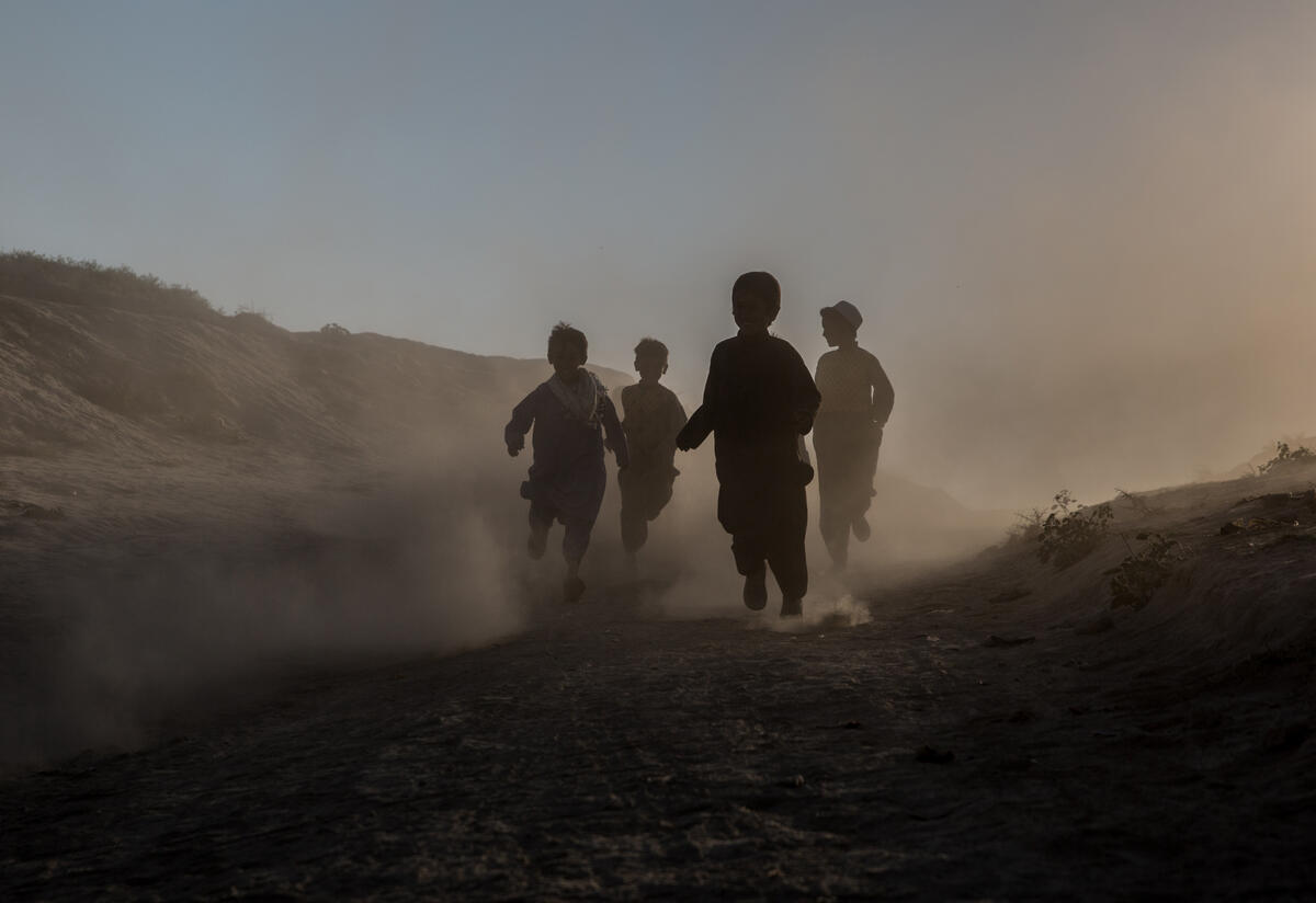 Children run through a camp in Afghanistan for people displaced by drought. Children run through a desert and mountain landscape. The light is such that their faces are obscured.