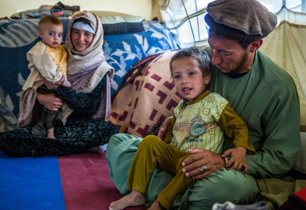 A mother and father in Afghanistan sit on the ground among blankets and pillows, each with a child on their lap. The boy in the front is smiling and his father is looking at him. 