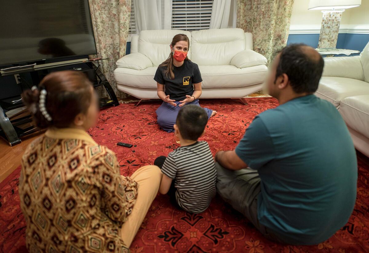 Jessica Carey, an IRC employment specialist, sits on the floor in a living room facing and talking to a family of three, parents and a young child.  
