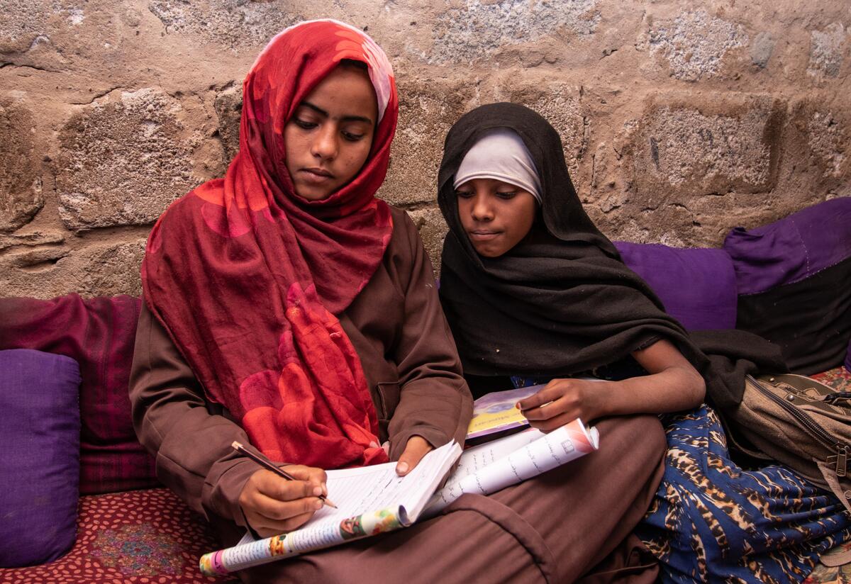 In a camp for displaced families in Yemen, Na'aem, 11 and Aisha, 10, sit on the ground going over a school workbook together and writing. The girls are best friends.