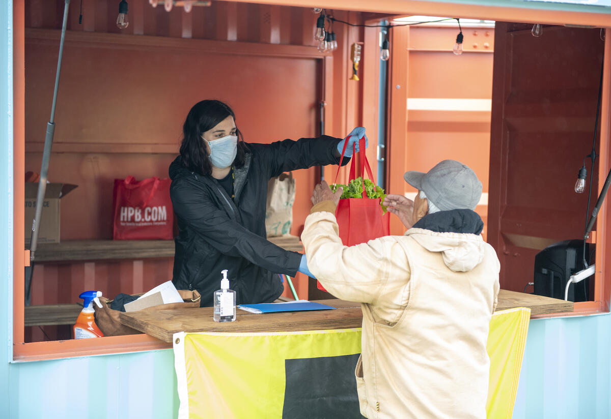 New Roots Coordinator delivers food to refugee families. A woman wearing a mask hands a tote bag of food to a refugee.