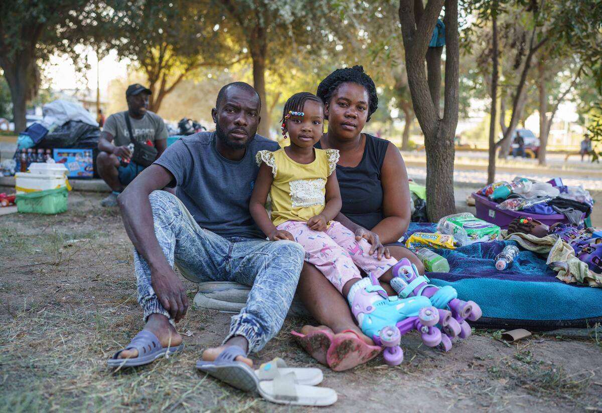 In a makeshift encampment in Mexico, a Haitian family--a mom, dad and young daughter--look straight at the camera while sitting on the ground next to their suitcases and blankets. 