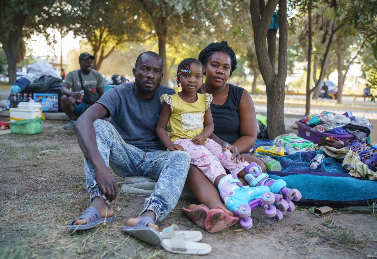 A Haitian family in a makeshift encampment in Mexico. The Biden Administration is using public health code Title 42 to turn away Haitian asylum seekers at the border. In a makeshift encampment in Mexico, a Haitian family--a mom, dad and young daughter--look straight at the camera while sitting on the ground next to their suitcases and blankets.