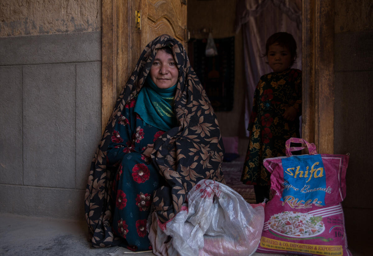 An Afghan mother sits next to large sack of rice with her toddler standing in the doorway behind her.