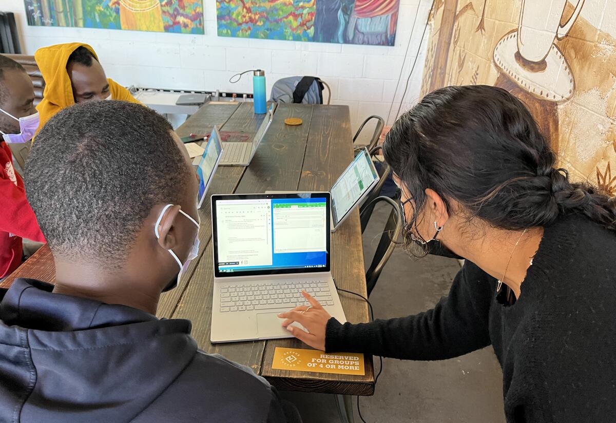 Resettled refugees complete Microsoft Excel training at the IRC Three students sitting at a table with their computers. Their teacher is leaning over to help one student at their computer.