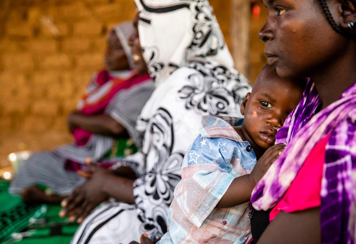A mother holds her baby while seated on the ground listening to an IRC malnutrition counseling session in Chad.