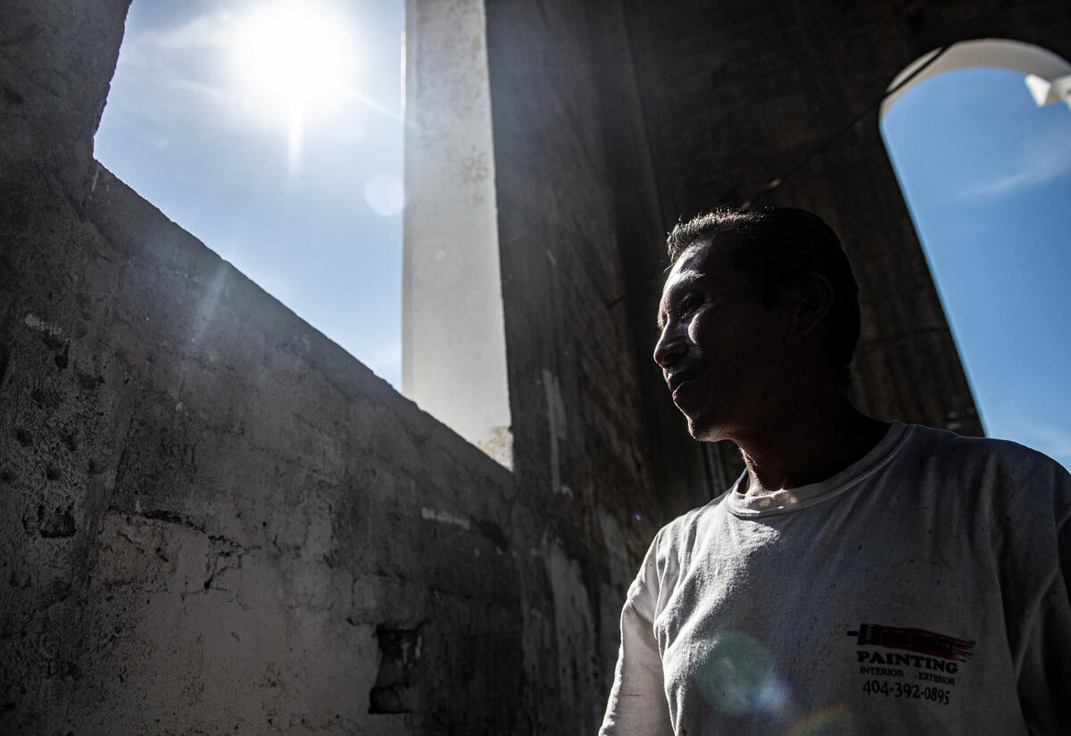 A Salvadaron man, Reynaldo, stands looking out at the sunshine through the window of a church he has been hired to paint.painting, 