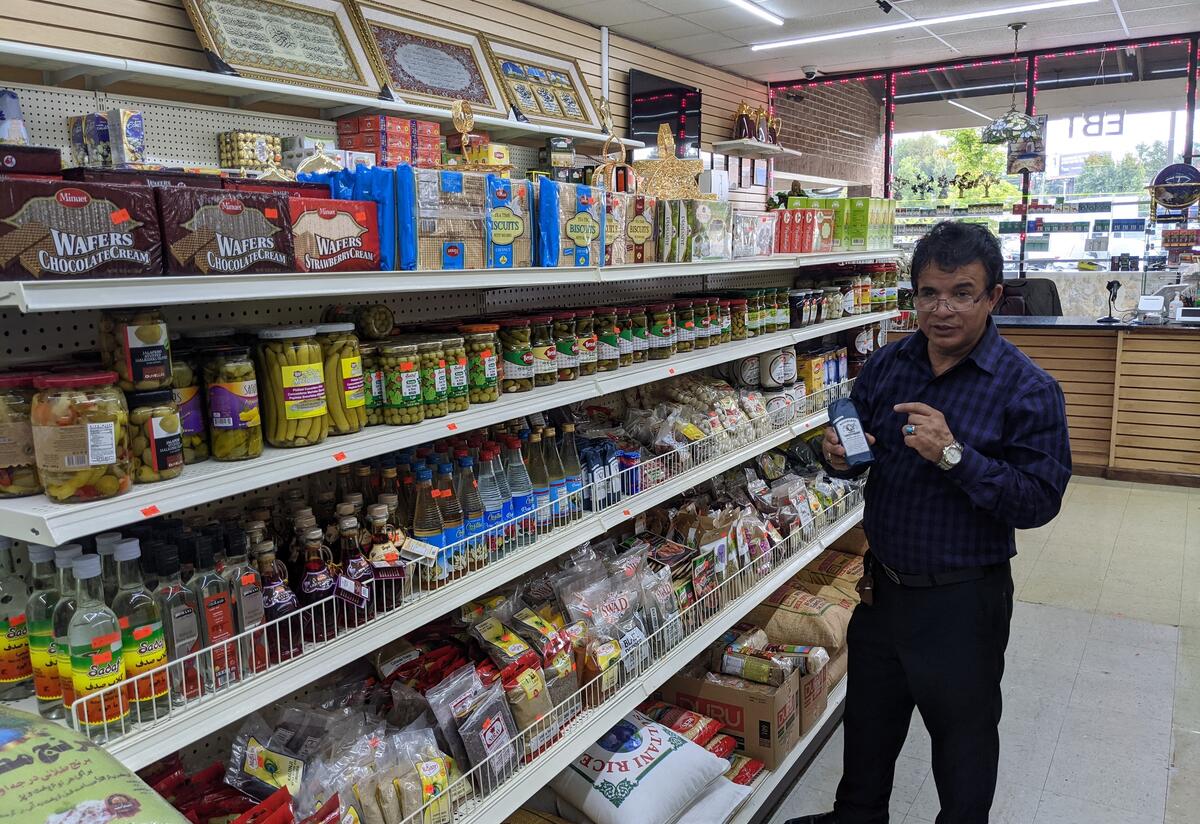 Storeowner standing next to shelf of goods including biscuits, wafers, and bags of rice.