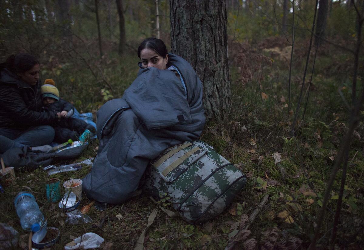 In a forest, a woman sits on the ground in a sleeping bag next to another woman and a child. They have backpacks and other supplies on the ground, and it is clearly cold outside. 