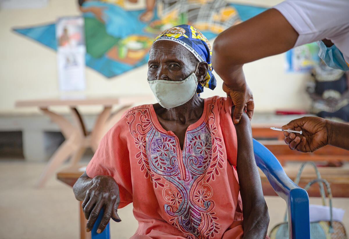 Enike Soduku, an elderly refugee, sits in a chair at an IRC clinic in Uganda as she receives a COVID-19 vaccination.
