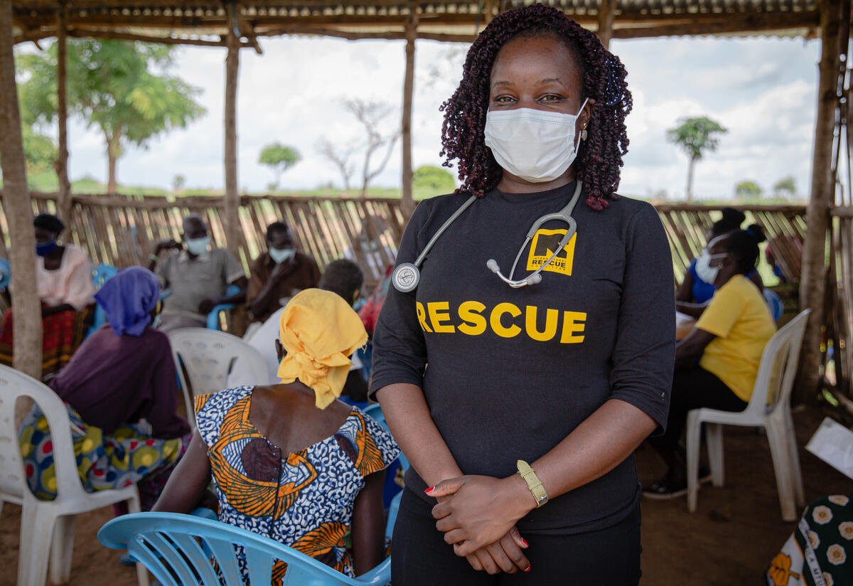 Dr. Ruth Grace Babirye with elderly refugees receiving vaccinations at an IRC center in Bidi Bidi, Uganda. Dr. Ruth Grace Babirye stands wearing a protective face mask as elderly people sit in an open waiting room in Bidi Bidi refugee settlement in Uganda to get vaccinated against COVID-19.