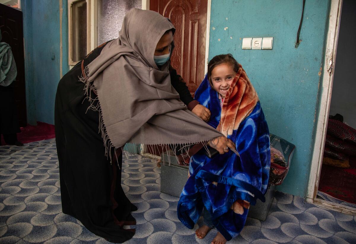 Noor wraps her daughter in a blanket purchased with emergency cash assistance provided by the IRC. Noor bends over to adjust a blue blanket her daughter has draped over her shoulders. Her daughter is smiling.