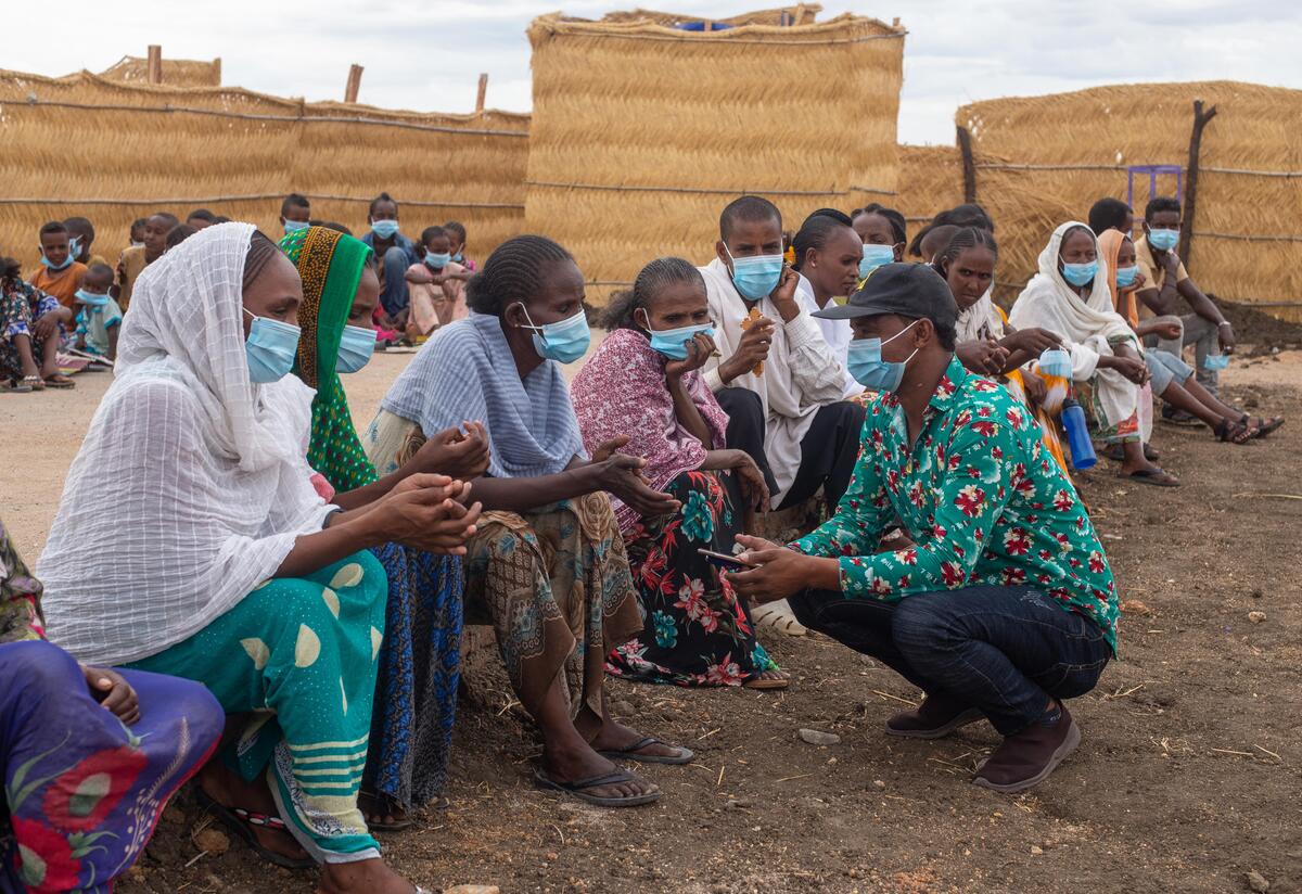People in Tunaydbah camp in Sudan sit on benches while one man kneels in front of them. 