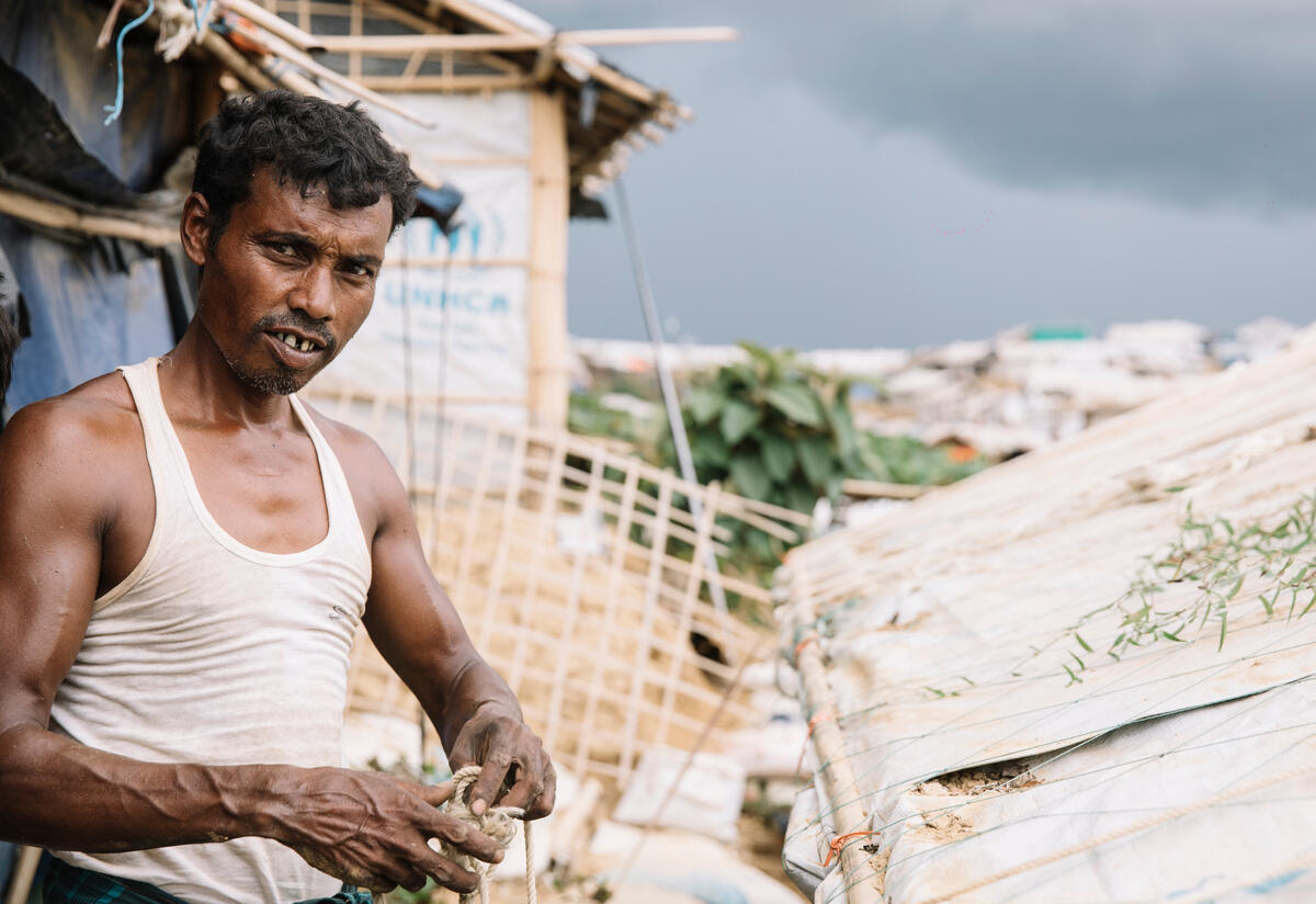 A man and his son rush to secure their roof as storm clouds gather over Kutupalong Refugee Camp in Bangladesh. A man looks at the camera while standing in front of a caved in roof.