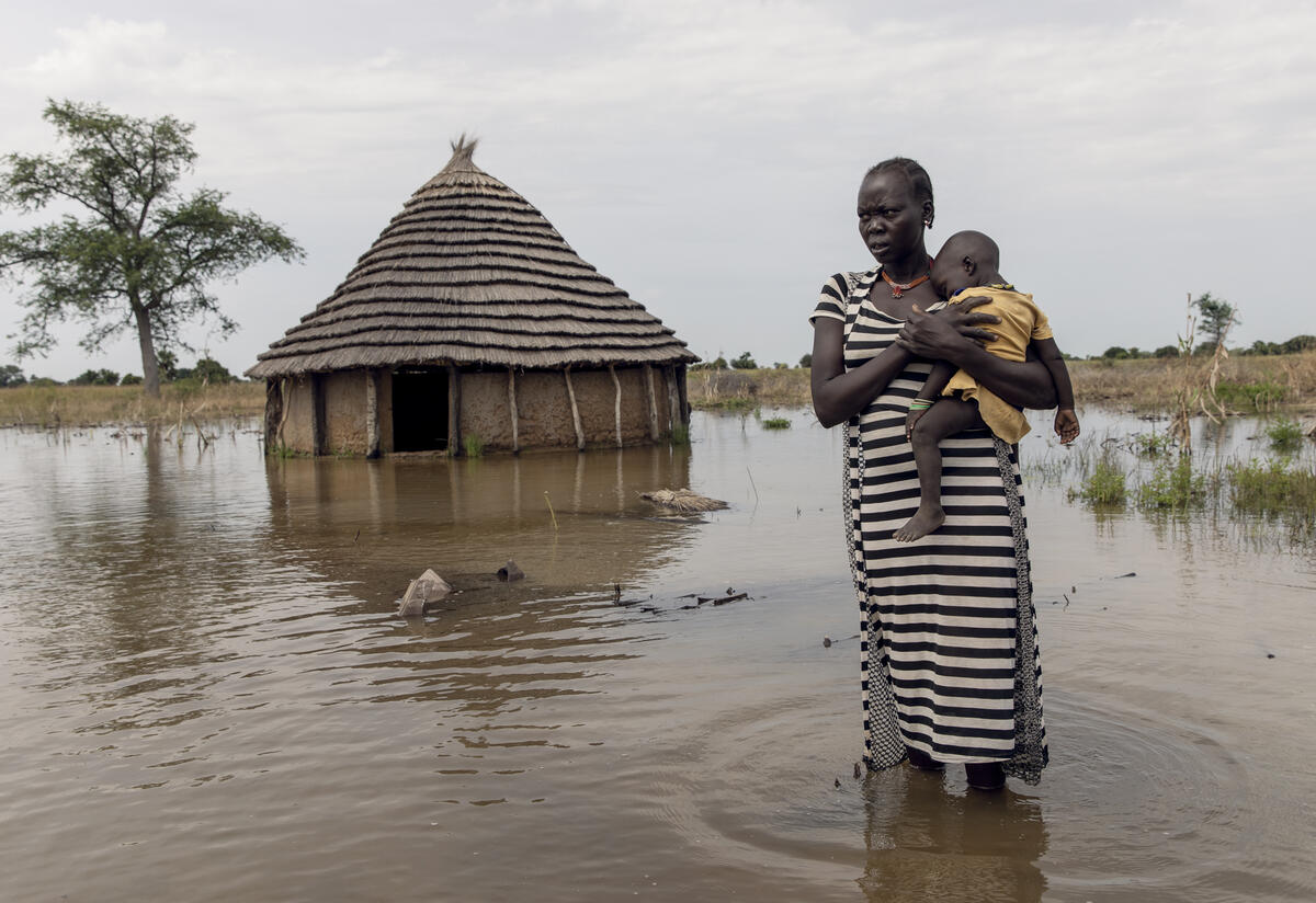 With water above her ankles, a mother holds her child and stands in front of her flooded home. 