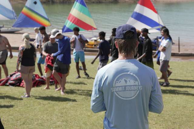 Man stands with his back turned in the foreground, wearing a Summit Journeys blue long-sleeve shirt and a blue ball cap. Background includes 10+ youth grabbing life jacket to prepare for a sailing activity on the lake shown in the far background. 