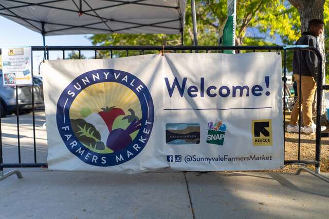 Sunnyvale Farmers Market vinyl sign hangs on a metal fencing barrier in front of a market canopy tent at Sunnyvale Park. The banner reads "Welcome!"