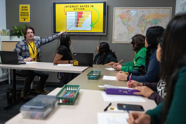 Will Weide, financial capabilities coordinator, sits to the left at a table pointing to a projected display of the financial literacy training. Five students sit at the table in the background/foreground of the photo.