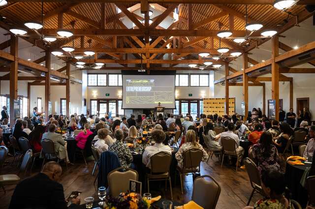 Seated guests captured inside The Garden Place, image from the back of the room looking forward to the stage and presentation screen. Guests are facing the screen seated at round tables in a vaulted room with wooden beams and rafters visible.