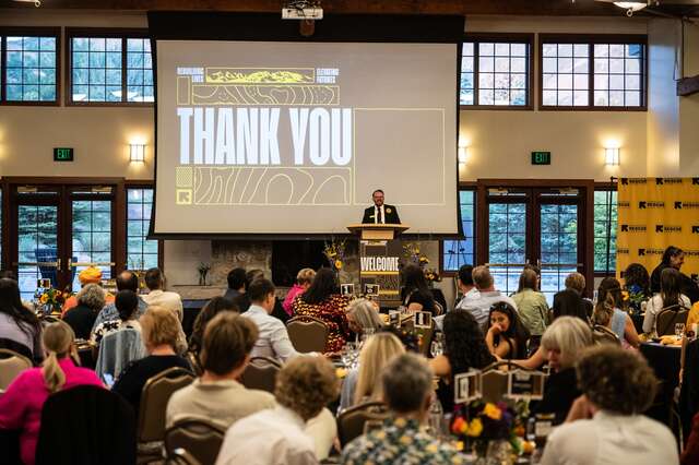 Danny Beus, executive director at the International Rescue Committee in Utah, thanks guests for attending from the podium, the screen behind him reads "Thank you." Seated guests look toward the podium, away from the camera.