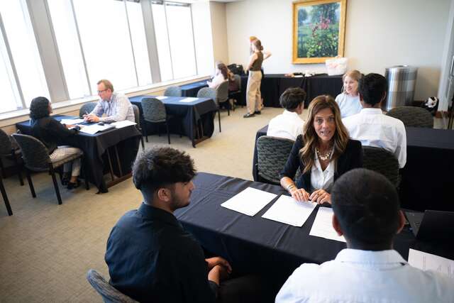 From the back of the room, youth participants are sitting across from Zions Bank recruiters--there are three tables with black tablecloths occupied by one or two participants and one recruiter as they complete mock interviews. A golde framed painting adorns the back wall, windows on the left light up the room.