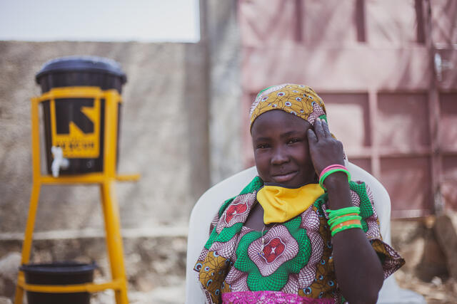 10-year old Anastasie sits in front of an IRC hand washing station and smiles at the camera.