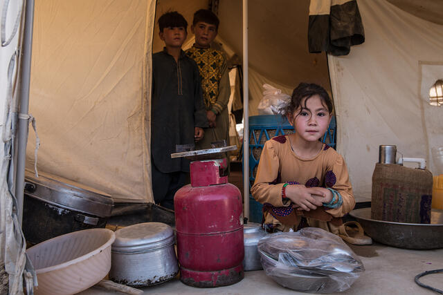 A young girl stands behind cooking pots and pans. Two boys stand in the doorway of a temporary shelter behind her.
