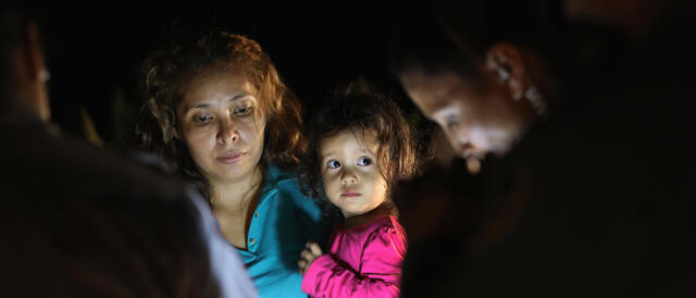A mother holding her young daughter claiming asylum with two border control agents.