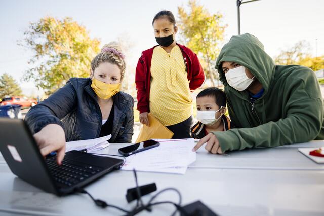 A family gathers with an educator outdoors around a laptop to complete activities for their students, all wear masks.