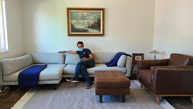 Salam sits on a large white couch in a freshly set up apartment, masked and looking at his phone with a smile.
